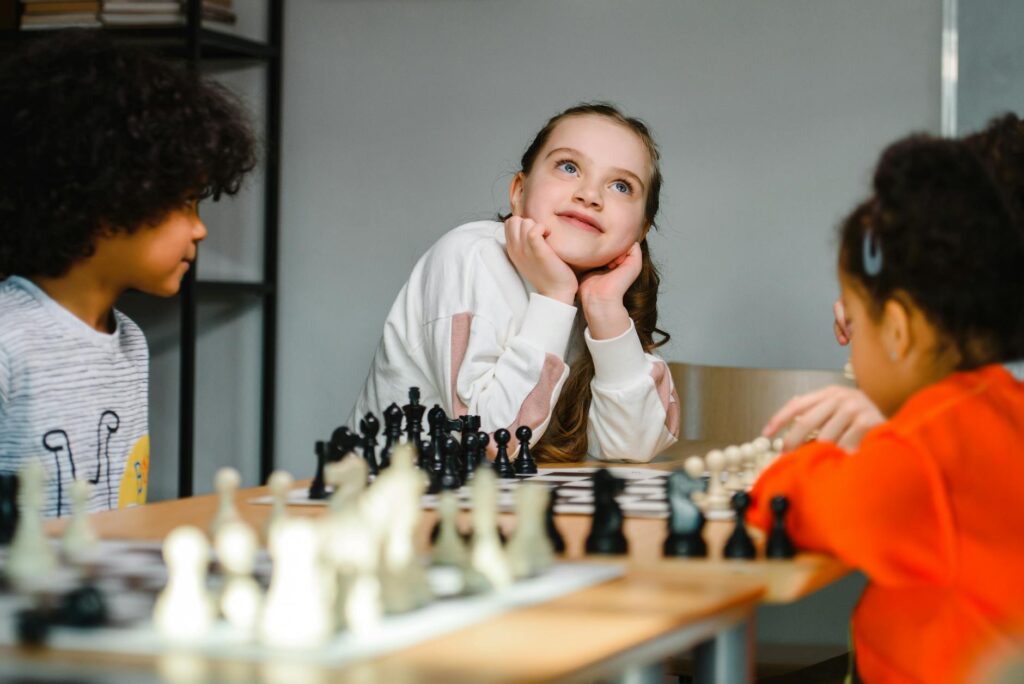 girl in white sweater sitting beside kids playing chess looking up