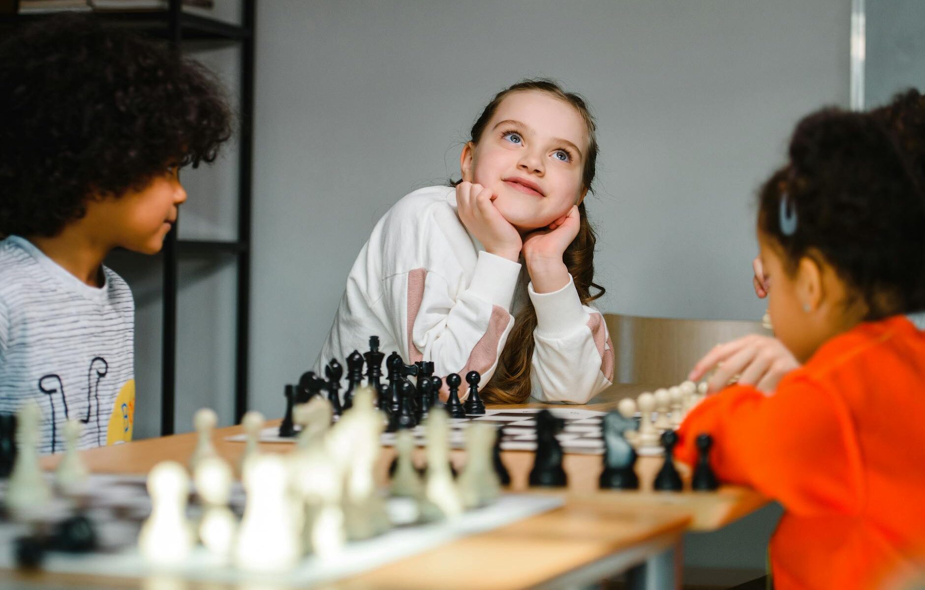 girl in white sweater sitting beside kids playing chess looking up